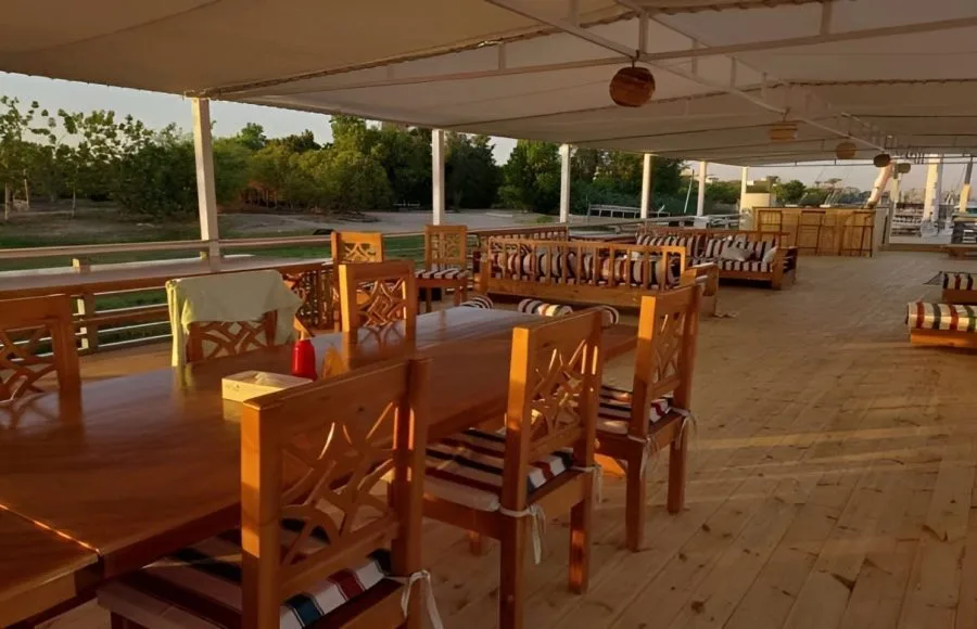 An outdoor dining deck on a cruise ship with wooden tables, chairs, and a view of the water and shore.