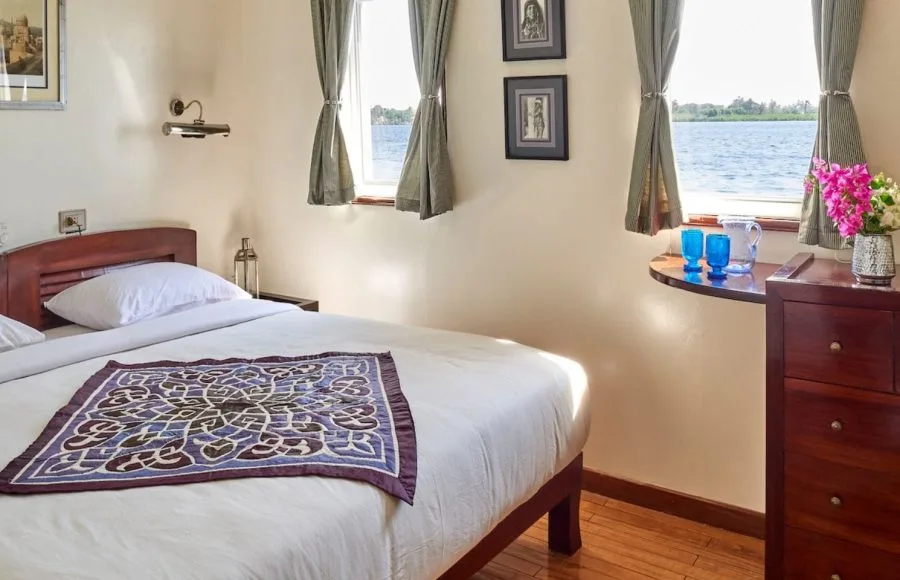 Interior view of a cabin on the Lazuli Albatros Dahabiya Nile Cruise, featuring a double bed, wooden furniture, and a window view of the Nile River.