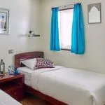 A cabin interior featuring two twin beds, a small nightstand, and blue curtains on the Lazuli Albatros Dahabiya Nile Cruise.