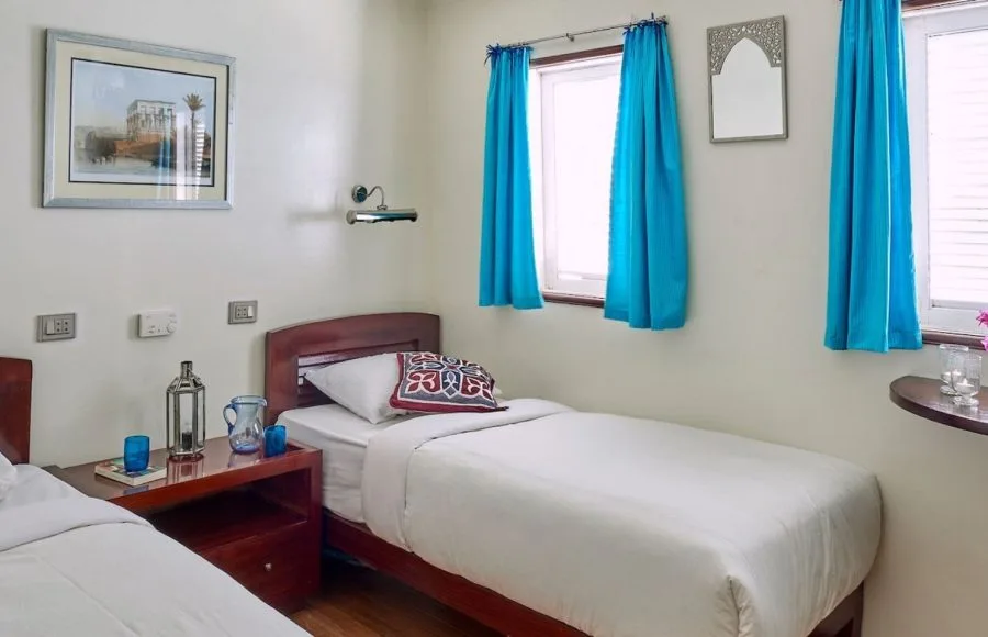 A cabin interior featuring two twin beds, a small nightstand, and blue curtains on the Lazuli Albatros Dahabiya Nile Cruise.