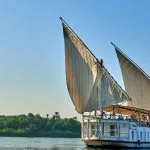 A traditional Egyptian two-masted wooden sailing boat (dahabiya) named Lazuli Albatros cruising on the Nile river with lush green banks in the background.