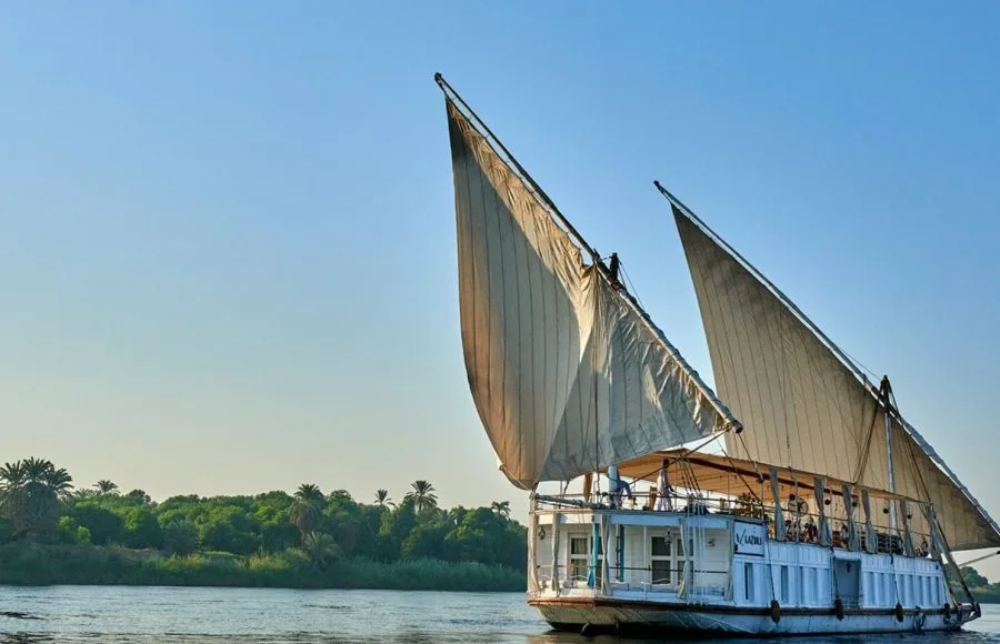A traditional Egyptian two-masted wooden sailing boat (dahabiya) named Lazuli Albatros cruising on the Nile river with lush green banks in the background.