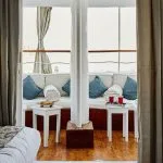 A cozy cabin bedroom interior aboard the Lazuli Roi Farouk Dahabiya Nile Cruise boat, featuring twin beds with patterned blankets, large windows with flowing curtains, and a small sitting area with white side tables and blue cushions