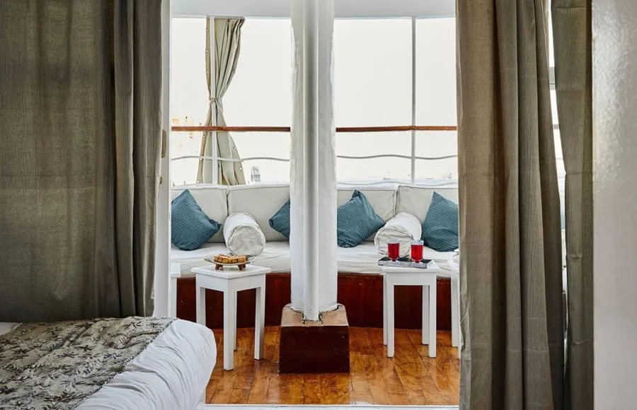 A cozy cabin bedroom interior aboard the Lazuli Roi Farouk Dahabiya Nile Cruise boat, featuring twin beds with patterned blankets, large windows with flowing curtains, and a small sitting area with white side tables and blue cushions