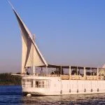 A traditional, two-masted white wooden dahabiya sailboat with large white sails gliding on the tranquil blue waters of the Nile River, with lush green riverbanks visible in the background.