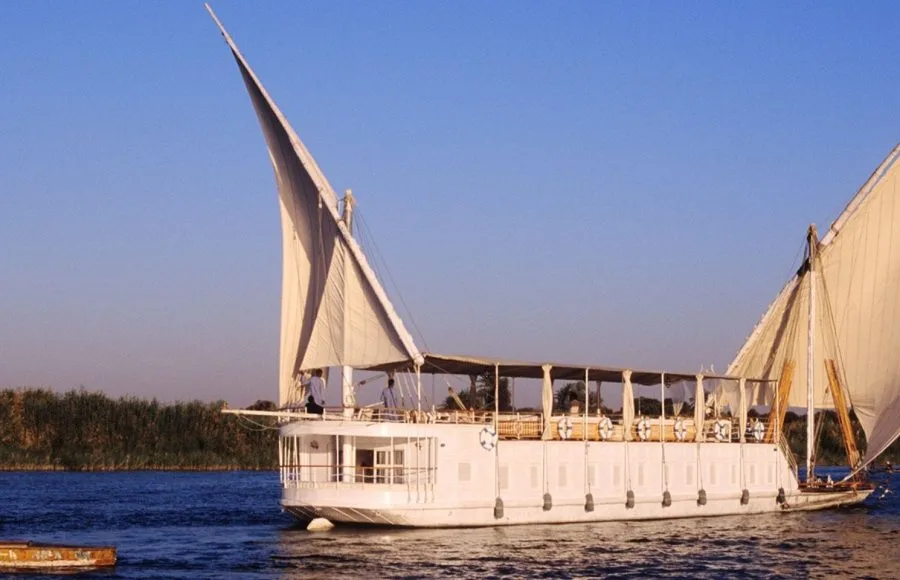 A traditional, two-masted white wooden dahabiya sailboat with large white sails gliding on the tranquil blue waters of the Nile River, with lush green riverbanks visible in the background.