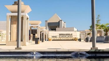 The modern entrance of the National Museum of Egyptian Civilization (NMEC) building with a water feature and clear blue sky.