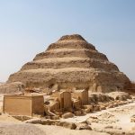 The large, tiered structure of the Saqqara Step Pyramid Of Pharaoh Djoser, partially under restoration scaffolding, visible in the desert of Saqqara, Egypt, with surrounding complex ruins in the foreground.