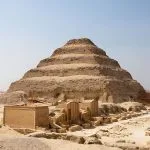 The large, tiered structure of the Saqqara Step Pyramid Of Pharaoh Djoser, partially under restoration scaffolding, visible in the desert of Saqqara, Egypt, with surrounding complex ruins in the foreground.