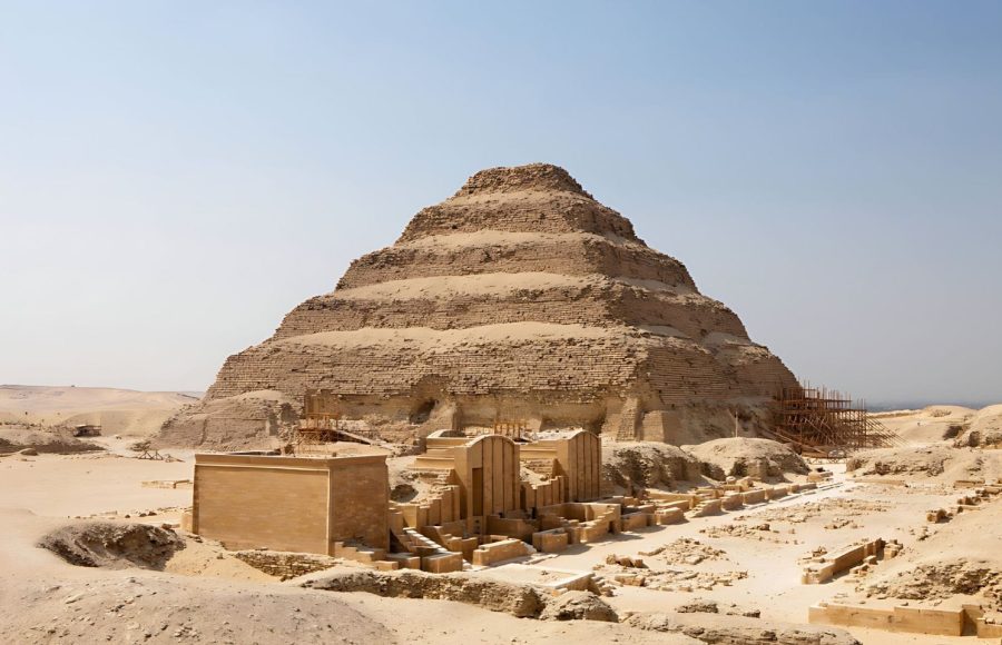 The large, tiered structure of the Saqqara Step Pyramid Of Pharaoh Djoser, partially under restoration scaffolding, visible in the desert of Saqqara, Egypt, with surrounding complex ruins in the foreground.