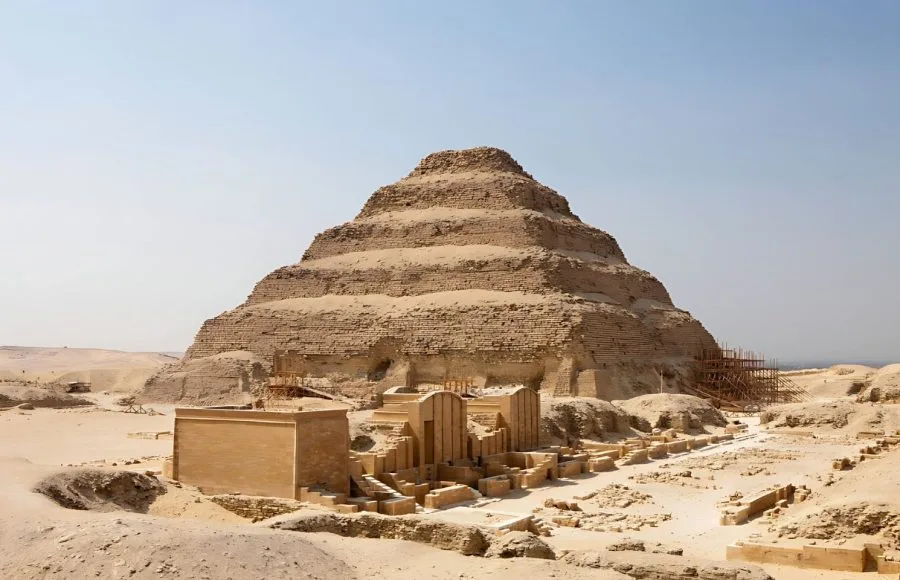 The large, tiered structure of the Saqqara Step Pyramid Of Pharaoh Djoser, partially under restoration scaffolding, visible in the desert of Saqqara, Egypt, with surrounding complex ruins in the foreground.