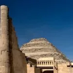An imposing view of the six-tiered The Step Pyramid Of Zoser in Saqqara, Egypt, framed by the ruins of a partially restored fluted column and stone wall from the complex.