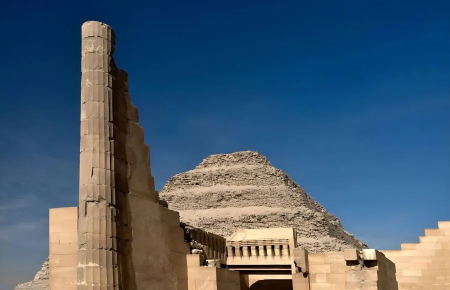 An imposing view of the six-tiered The Step Pyramid Of Zoser in Saqqara, Egypt, framed by the ruins of a partially restored fluted column and stone wall from the complex.