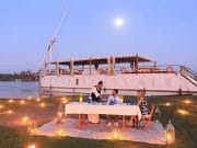 A couple enjoys a romantic dinner lit by lanterns on the grassy bank beside a traditional Dahabiya sailing boat under a full moon, perfect for those looking to cruise the Nile on a Dahabiya or enjoy a Dahabiya Nile Cruise from Aswan to Luxor.