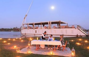 A couple enjoys a romantic dinner lit by lanterns on the grassy bank beside a traditional Dahabiya sailing boat under a full moon, perfect for those looking to cruise the Nile on a Dahabiya or enjoy a Dahabiya Nile Cruise from Aswan to Luxor.