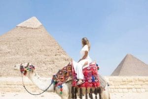 A woman in a white outfit rides a colorfully adorned camel, looking towards the majestic Great Pyramids of Giza under a clear blue sky in Egypt.