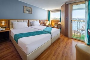 A comfortable cabin on a standard Nile cruise ship with two twin beds, a balcony overlooking the Nile, and simple furnishings.