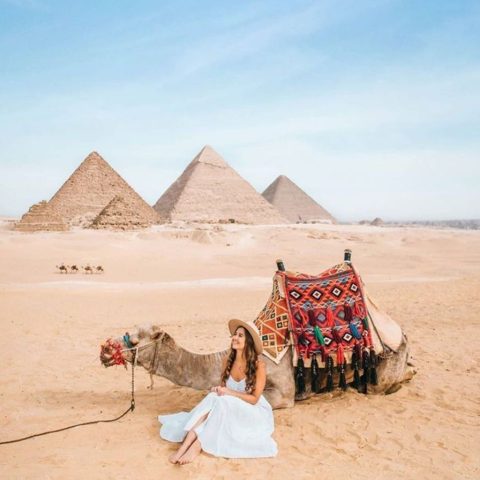 A tourist in a white dress sitting next to a camel with the Pyramids of Giza in the background, perfect for 7 day Egypt tour packages.