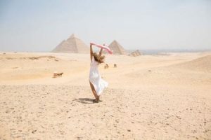 A joyful family, with a father giving a piggyback ride to his daughter while his wife laughs beside them, stands on a sandy beach at the edge of the clear blue sea under a bright, sunny sky, reflecting a relaxing part of a long Egypt itinerary.