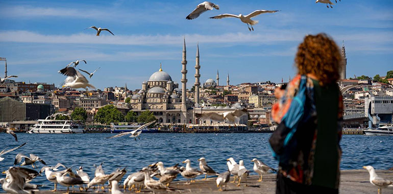 A woman watches seagulls in flight on the edge of the Bosphorus with the Yeni Cami Mosque and Istanbul in the background