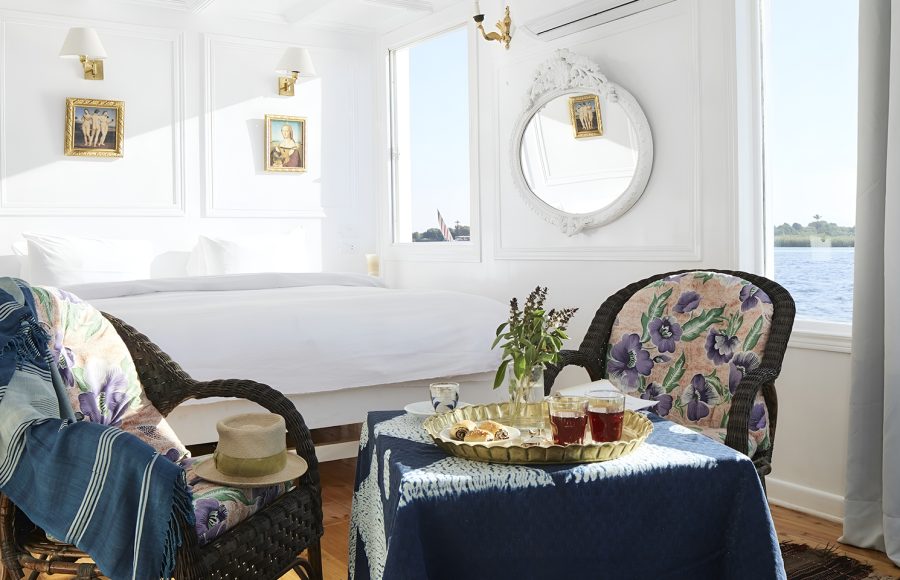 A cozy seating area in a Nile cruise cabin with wicker chairs, a blue draped table, and large windows overlooking the river.