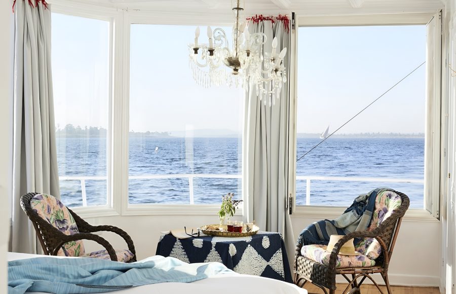 Sunlit cabin interior of a Nile cruise boat featuring wicker chairs and a large bay window overlooking the river.