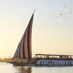 A traditional Egyptian Dahabiya sailing boat with red and white striped sails on the Nile River at sunset.