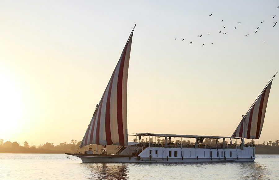 A traditional Egyptian Dahabiya sailing boat with red and white striped sails on the Nile River at sunset.