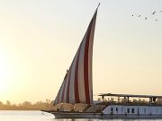 A traditional Egyptian Dahabiya sailing boat with red and white striped sails on the Nile River at sunset.