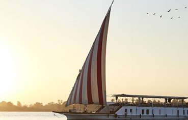 A traditional Egyptian Dahabiya sailing boat with red and white striped sails on the Nile River at sunset.