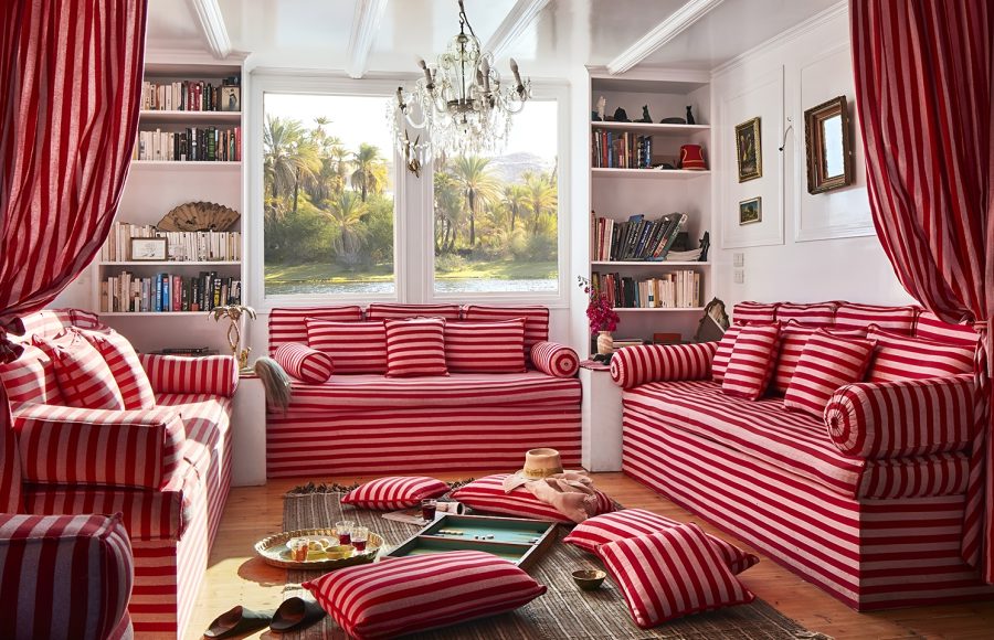 A cozy lounge on an Egyptian dahabiya boat featuring red and white striped sofas and floor cushions, with a view of the Nile through a large window.