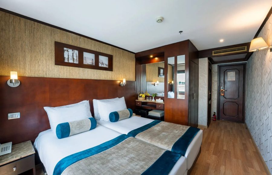 Comfortable twin beds in a cabin on the Nile Admiral cruise ship with wood flooring, classic Egyptian decor, and a vanity area.