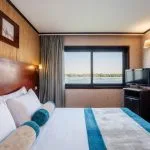 Nile Admiral cruise ship cabin with a large bed, TV, and a window offering a scenic view of the Nile River and the green riverbanks.