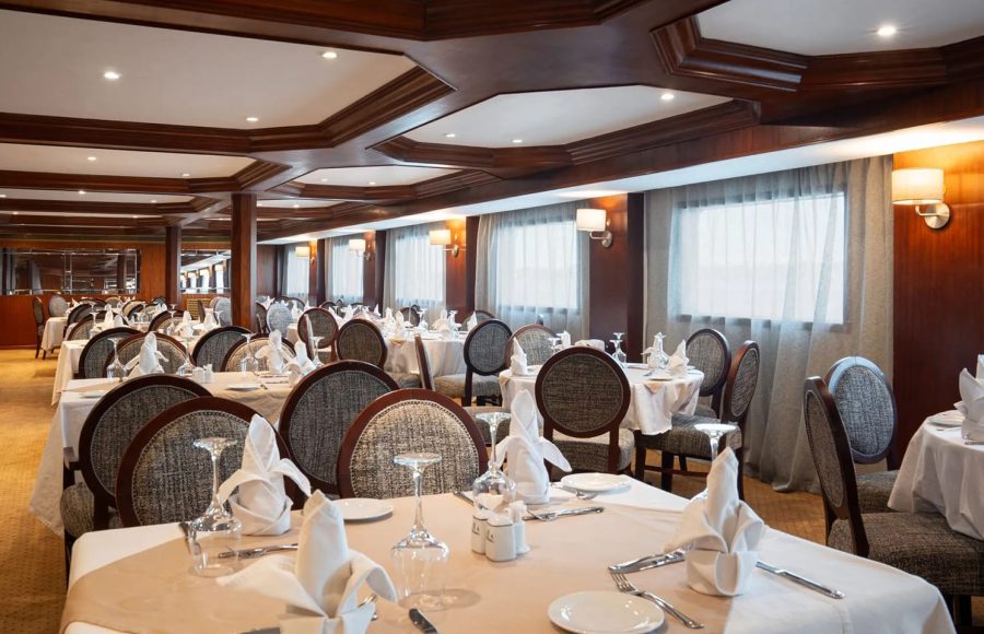 Wide view of the elegantly carpeted main dining room on the Nile Admiral cruise ship with set tables, upholstered chairs, and wood-paneled ceilings.