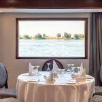Close-up of a formally set round dining table for four, with a large window in the background showing the Nile River and palm trees.