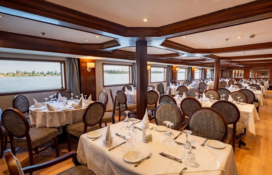 Formal dining room on the Nile Admiral cruise ship with set tables, elegant chairs, dark wood trim, and large windows viewing the Nile River.