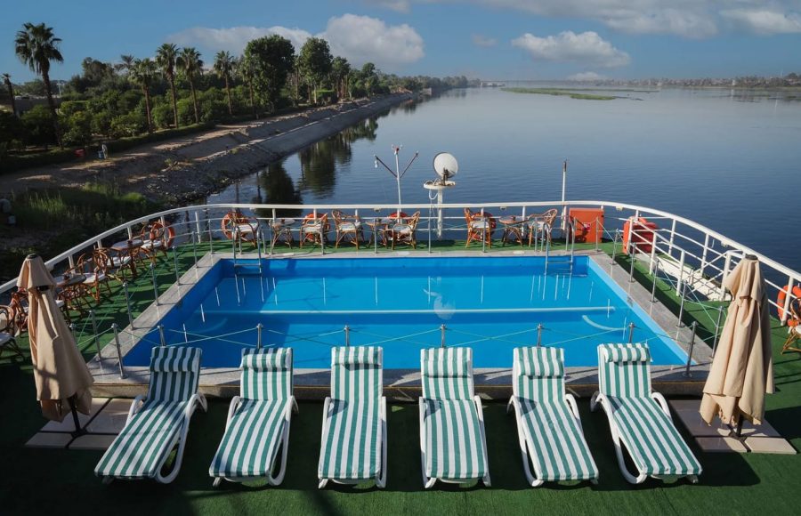 Swimming pool and sun loungers on the upper deck of the Nile Admiral cruise ship, offering a panoramic view of the Nile River and palm-lined banks.