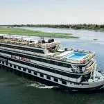 Nile cruise ship "Nile Admiral" sailing on the Nile River under a clear sky, featuring an upper deck swimming pool and sun loungers.