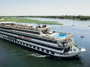 Nile cruise ship "Nile Admiral" sailing on the Nile River under a clear sky, featuring an upper deck swimming pool and sun loungers.