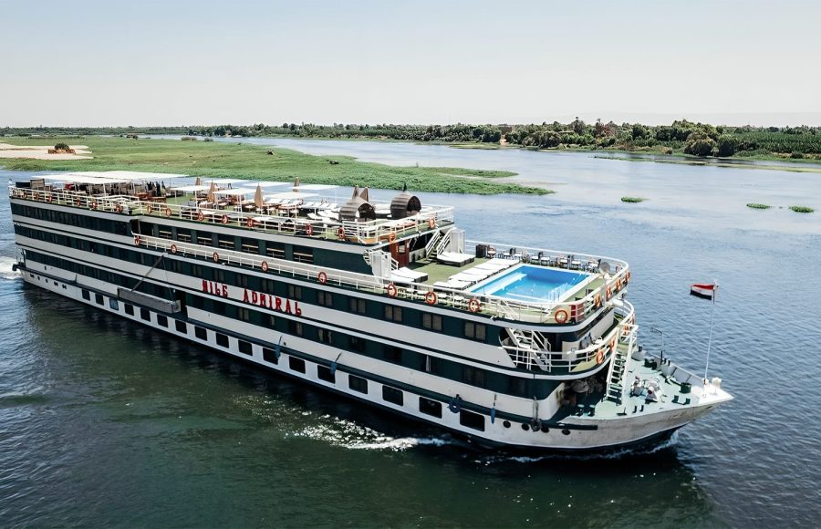 Nile cruise ship "Nile Admiral" sailing on the Nile River under a clear sky, featuring an upper deck swimming pool and sun loungers.