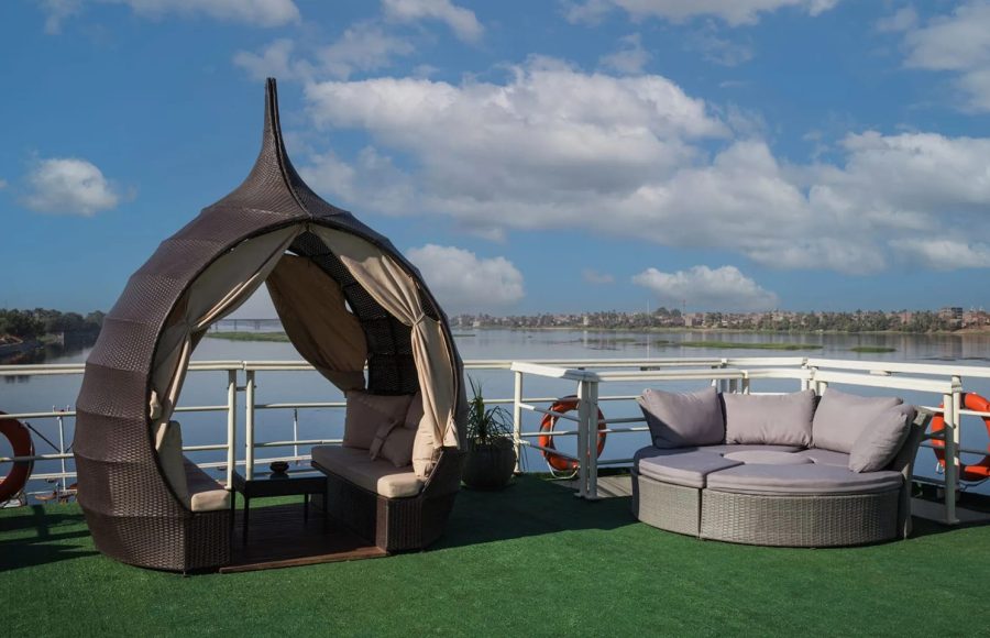 Sun deck of the Nile Admiral cruise ship with a wicker cabana and round sun lounger, overlooking the Nile River under a blue sky.