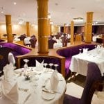 The formal dining room of the Al-Hambra Nile Cruise with white tablecloths, purple velvet booth seating, and gold-colored pillars. The tables are set with fine glassware and white napkins in an elegant, well-lit space.