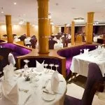 The formal dining room of the Al-Hambra Nile Cruise with white tablecloths, purple velvet booth seating, and gold-colored pillars. The tables are set with fine glassware and white napkins in an elegant, well-lit space.