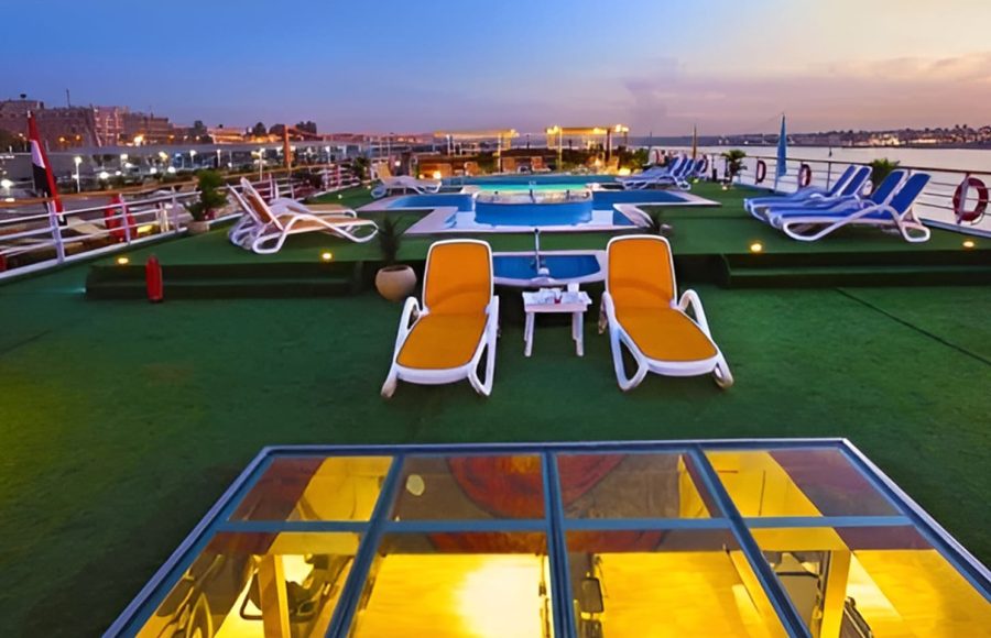 The outdoor sun deck of the Al-Hambra Nile Cruise at dusk, featuring green artificial turf, yellow and white lounge chairs, a small swimming pool, and glass skylights looking into the lower decks.