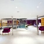 A modern and spacious lounge on the Al-Hambra Nile Cruise with white floors, purple velvet chairs, and silver support pillars. A glass spiral staircase serves as a focal point in the center, with a wooden reception desk visible in the background.