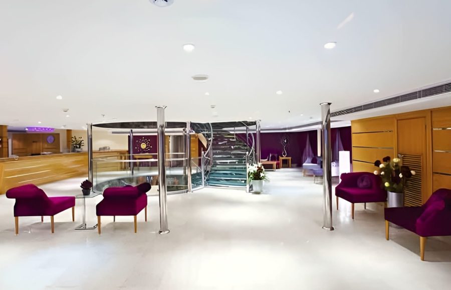 A modern and spacious lounge on the Al-Hambra Nile Cruise with white floors, purple velvet chairs, and silver support pillars. A glass spiral staircase serves as a focal point in the center, with a wooden reception desk visible in the background.