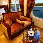 A well-lit luxury cabin interior on the Al-Hambra Nile Cruise featuring a bronze-textured sofa, a bed with white linens, and large panoramic windows overlooking the Nile River and green shoreline. A glass coffee table with wine glasses sits on a circular rug in the foreground.