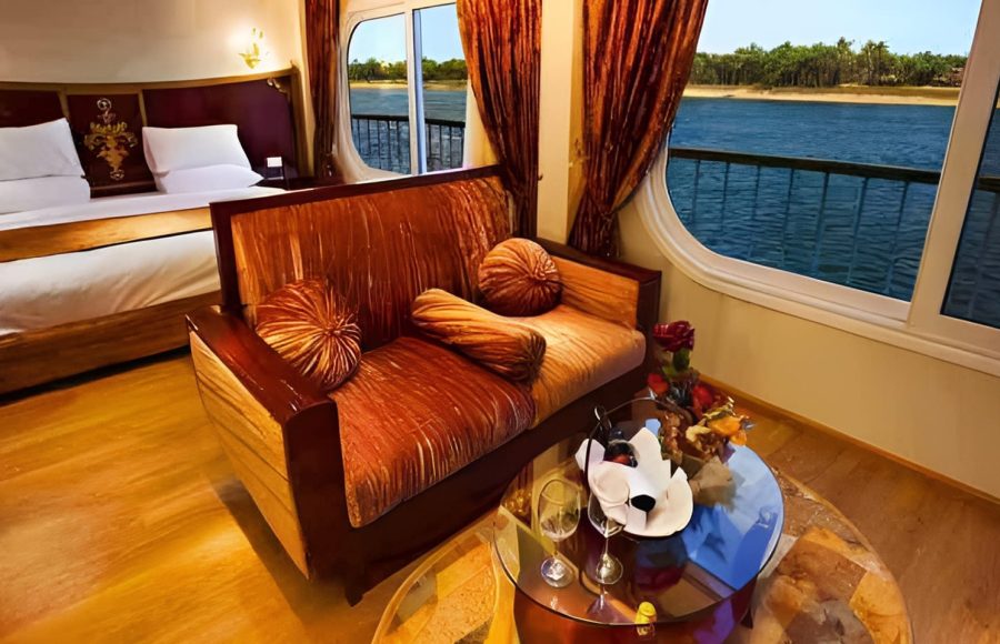 A well-lit luxury cabin interior on the Al-Hambra Nile Cruise featuring a bronze-textured sofa, a bed with white linens, and large panoramic windows overlooking the Nile River and green shoreline. A glass coffee table with wine glasses sits on a circular rug in the foreground.