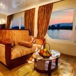 A luxury suite on the Al-Hambra Nile Cruise featuring a gold-textured sofa, a glass coffee table with a fruit basket, and a plush bed. Large panoramic windows offer a stunning view of the Nile River and distant mountains during a vibrant orange and pink sunset.
