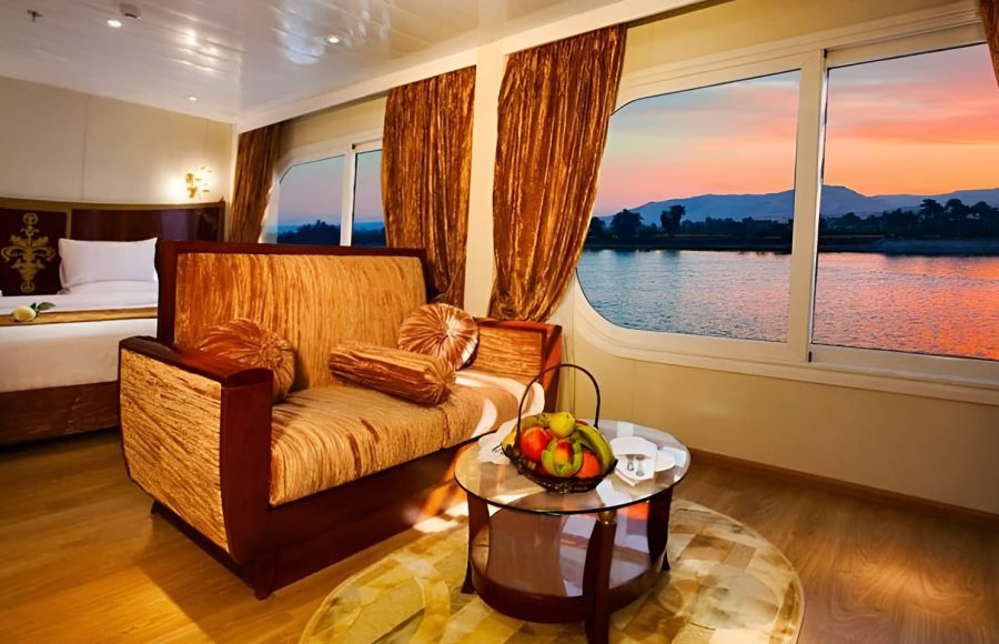 A luxury suite on the Al-Hambra Nile Cruise featuring a gold-textured sofa, a glass coffee table with a fruit basket, and a plush bed. Large panoramic windows offer a stunning view of the Nile River and distant mountains during a vibrant orange and pink sunset.